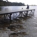 Persona sentada al final de un muelle plástico Maderplast sobre cuerpo de agua, en un entorno natural y tranquilo.